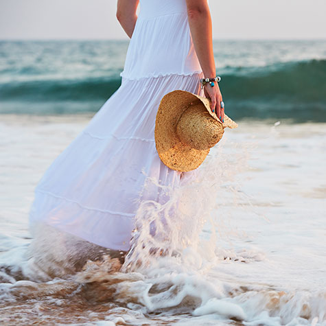 Woman walking in the surf as the sun goes down, holding a straw hat